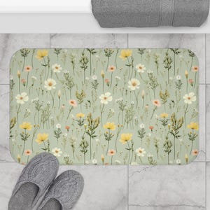 May include: A green bath mat with a floral pattern featuring white, yellow, and pink flowers. The flowers are scattered across the mat, creating a whimsical and delicate design.