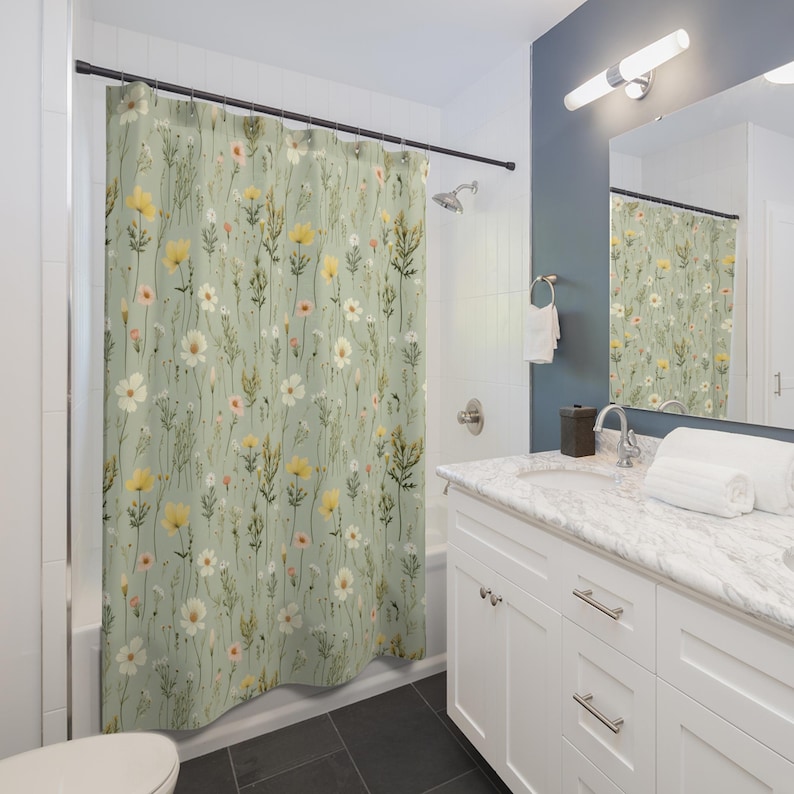 May include: A shower curtain with a floral pattern featuring yellow, white, and pink wildflowers on a light green background. The curtain is hanging in a bathroom with white cabinets and a marble countertop.