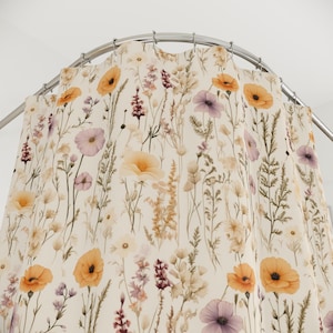 May include: A floral shower curtain featuring a cream background with a pattern of wildflowers in shades of orange, purple, and yellow. The curtain is hanging from a curved, silver-colored shower rod. The design evokes a natural, botanical theme.