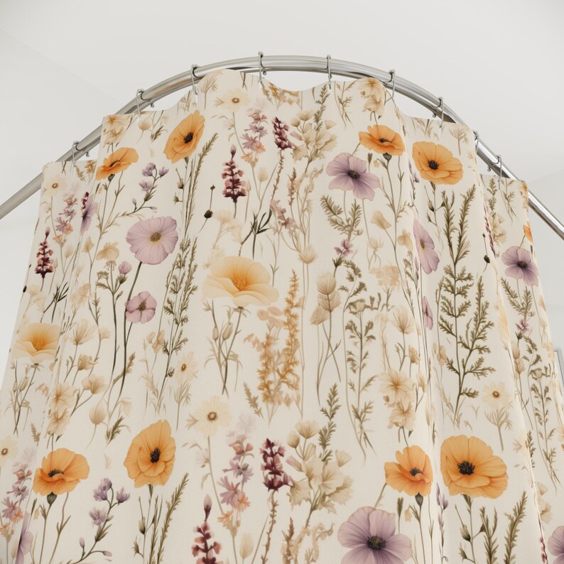 May include: A floral shower curtain featuring a cream background with a pattern of wildflowers in shades of orange, purple, and yellow. The curtain is hanging from a curved, silver-colored shower rod. The design evokes a natural, botanical theme.
