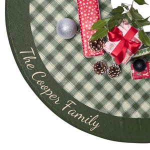 May include: A green and white plaid Christmas tree skirt with the text "The Cooper Family" in white script. The skirt is decorated with wrapped gifts, pinecones, and a silver ornament. A red ribbon adorns one of the gifts.