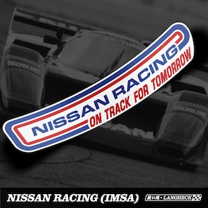 Op de afbeelding: Een witte, rode en blauwe sticker met de tekst "NISSAN RACING ON TRACK FOR TOMORROW" in een gebogen ontwerp. De sticker is geplaatst tegen een wazige afbeelding van een raceauto.