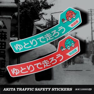 Puede incluir: Dos pegatinas de seguridad vial de Akita. Una es verde azulado con una tortuga roja, la otra es roja con una tortuga verde azulado. Ambas tienen texto japonés y el sello de la prefectura de Akita. Las pegatinas son curvas.