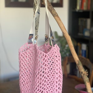 May include: A light pink crocheted tote bag with white straps featuring a marbled pattern. The bag is suspended from a wooden branch. The open weave design of the bag is visible, showcasing its handmade quality.