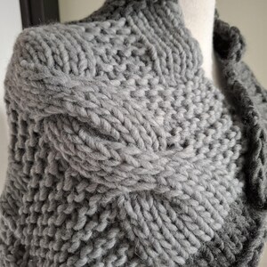 May include: A grey and black knitted cowl with a cable knit pattern. The cowl is made of a soft, chunky yarn.
