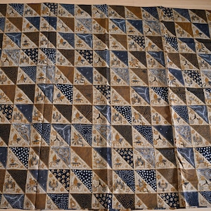 Puede incluir: Textil estampado con un diseño geométrico repetitivo. La tela presenta una cuadrícula de triángulos en tonos marrón, azul marino y crema. Algunos triángulos tienen patrones de puntos o motivos florales. El textil se utiliza probablemente para ropa o decoración del hogar.