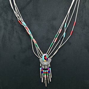 Puede incluir: Un collar de plata con un colgante ovalado central con un diseño de mosaico en rojo, azul, verde y amarillo. El collar tiene múltiples hilos con cuentas de colores y barras colgantes de plata, sobre un fondo negro.