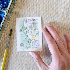 Puede incluir: Un mapa de acuarela dibujado a mano de la Tierra Media, una tierra ficticia de los libros de El Se&ntilde;or de los Anillos. El mapa muestra varios lugares, incluyendo la Comarca, Mordor y las Monta&ntilde;as Nubladas. El mapa est&aacute; dibujado en un estilo caprichoso con colores verde, marr&oacute;n y azul.