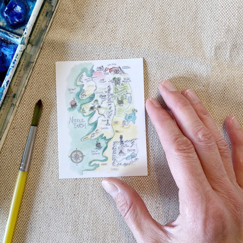 Puede incluir: Un mapa de acuarela dibujado a mano de la Tierra Media, una tierra ficticia de los libros de El Se&ntilde;or de los Anillos. El mapa muestra varios lugares, incluyendo la Comarca, Mordor y las Monta&ntilde;as Nubladas. El mapa est&aacute; dibujado en un estilo caprichoso con colores verde, marr&oacute;n y azul.