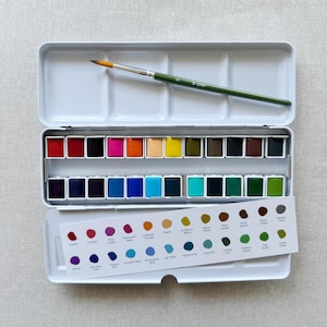 Puede incluir: Un set de acuarelas en caja de metal blanco con dos filas de veinticuatro pastillas rectangulares de colores. Un pincel verde descansa sobre la tapa abierta. Se incluye una carta de colores blanca con etiquetas impresas.