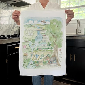 May include: A white tea towel with a watercolor illustration of the map from the children's book "The Tale of Peter Rabbit". The map shows the locations of Mr. McGregor's garden, the rabbit hole, and other landmarks from the story.