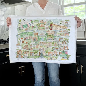 May include: A white tea towel with a watercolor illustration of the Florida State University campus. The illustration includes buildings, trees, and the text "Go 'Noles Florida State".