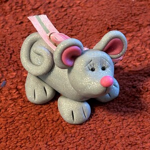 May include: A small, gray, glittery mouse figurine with pink ears and a pink nose. The mouse is wearing a pink and green ribbon around its neck.