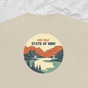 May include: A beige t-shirt with a circular graphic on the back. The graphic features a landscape scene with mountains, a lake, trees, and a person throwing a disc. The text "DISC GOLF STATE OF MIND" is above the graphic.