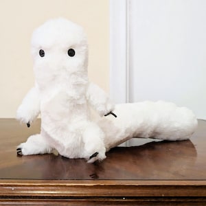 Puede incluir: Un peluche blanco con forma de criatura con cuerpo esponjoso, brazos pequeños y cola larga. Tiene ojos negros y pequeñas garras negras. El juguete está sobre una superficie de madera oscura.