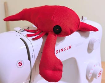 May include: A red fabric lobster toy with a black button eye, sitting on a white sewing machine with the brand name "SINGER" printed on it.