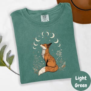 May include: A light green t-shirt with a graphic of a fox sitting under a crescent moon with stars. The fox is orange and white with a black nose and eyes. The text "COMFORT COLORS" is printed on the tag.