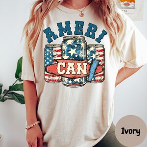 May include: A white t-shirt with a graphic of three red, white, and blue cans with the text "Ameri Can" printed on the front. The shirt is available in the color Ivory.