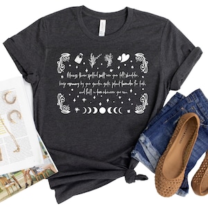 May include: A dark heather grey t-shirt with a white graphic design featuring a quote about throwing salt over your left shoulder, keeping rosemary by your garden gate, planting lavender for luck, and falling in love whenever you can. The graphic also includes a moon phase design and other botanical illustrations.