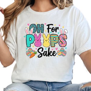 May include: White t-shirt with the phrase "OH For Peeps Sake" in colorful letters. The design includes Easter eggs, bunny ears, and a carrot. The letters are in various colors and patterns, creating a festive look.