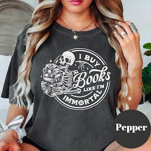 May include: A black t-shirt with a white graphic of a skeleton holding a stack of books. The graphic is surrounded by a circle with the text "I Buy Books Like I'm Immortal".