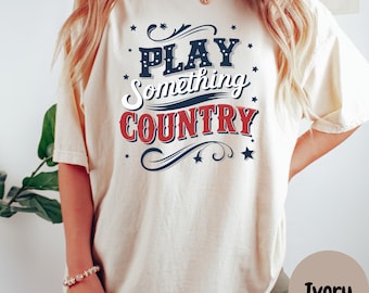 Puede incluir: Una camiseta blanca con un gráfico azul y rojo que dice "Play Something Country". La camiseta es de corte holgado y tiene cuello redondo.