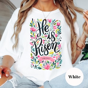 May include: White t-shirt featuring the text "He is Risen" in a decorative script, surrounded by a floral design with pink, yellow, and green elements. The text "Luke 24:6-7" is also present.