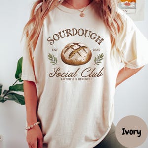 May include: A white t-shirt with the text "SOURDOUGH SOCIAL CLUB" in brown lettering. The text is surrounded by a brown circle with the words "EST. 2025" and a drawing of a loaf of sourdough bread. The shirt is labeled "Ivory".