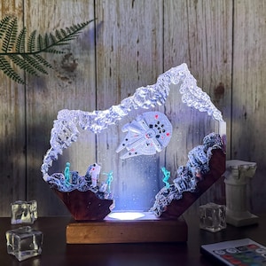 May include: A decorative light featuring a spaceship design with figures and a mountain-like structure. The spaceship is white and gray, with small figures in green and white. The base is brown, and the light has a wooden base.