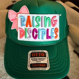 Può includere: Un cappellino trucker verde con una toppa rettangolare bianca che riporta il testo ricamato RAISING DISCIPLES in lettere multicolori. Un fiocco rosa è fissato sul lato sinistro della toppa. Un adesivo sulla visiera recita OTTO WORLD FAMOUS FOAM FRONT OSFM TRUCKER.