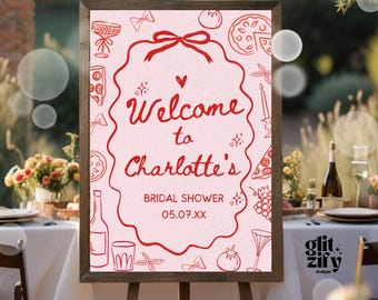 May include: A framed sign with a pink background and red text that reads "Welcome to Charlotte's Bridal Shower". The sign is decorated with red illustrations of food and drinks. The sign is displayed on a wooden easel.