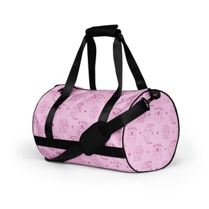 May include: A pink duffel bag with a repeating pattern of celestial symbols, including moons, stars, and eyes. The bag has black handles and a shoulder strap. The bag is designed for carrying items.