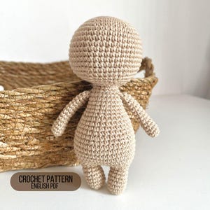 May include: A crocheted doll with a beige body and a round head. The doll is standing on a white surface next to a wicker basket.