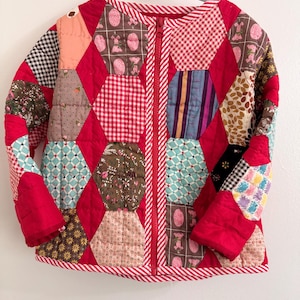 May include: A quilted jacket with a red background and a variety of patterned patches in shades of pink, brown, blue, and white. The jacket has a full zipper and is trimmed with red and white striped fabric. The sleeves are solid red.