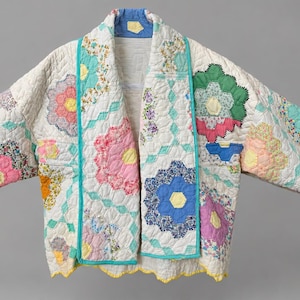May include: A quilted jacket featuring a white base with a multi-colored hexagonal flower pattern. The garment has long sleeves, a shawl collar, and a scalloped hemline finished with yellow trim. The floral motifs include pink, blue, and green.