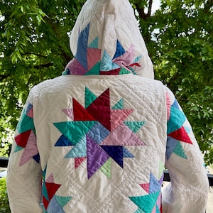 May include: A quilted jacket with a hood, featuring a starburst pattern on the back. The jacket is primarily white with colorful geometric shapes in shades of red, teal, purple, and blue. The hood is also white with similar colorful accents.