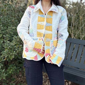 May include: A quilted jacket with a patchwork design. The jacket features a white base with yellow, red, and blue fabric squares. The jacket has a collar, pockets, and long sleeves. The jacket is worn over a black top and black pants.