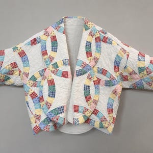 Puede incluir: Una chaqueta acolchada con base blanca y un colorido patrón de anillos de boda entrelazados. La prenda tiene mangas largas, frente abierto y una superficie texturizada. El diseño de patchwork incorpora varios estampados florales y geométricos.
