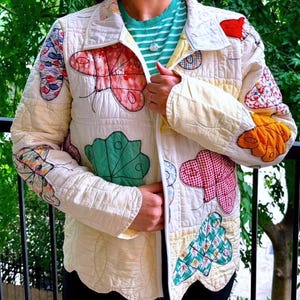 May include: A cream-colored quilted jacket featuring a variety of colorful butterfly-shaped fabric appliqués in shades of red, green, orange, and blue. The jacket has a collared neckline, a front zipper closure, and long sleeves with a grid-like stitched pattern.