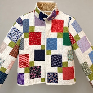 May include: A quilted jacket featuring a patchwork design of various colorful fabric squares in red, blue, green, and floral patterns. The garment has a high collar, long sleeves, and a light-colored quilted interior lining visible at the neck.