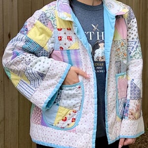 May include: A patchwork jacket with a variety of fabric patterns in pastel colors, including yellow, pink, and blue. The jacket has a light blue trim and pockets. The jacket is quilted and appears to be handmade.