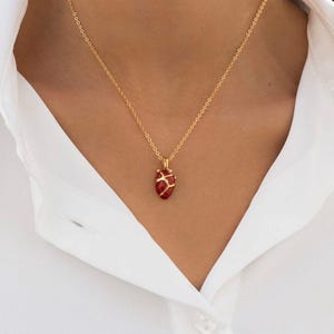 May include: A gold-colored necklace with a pendant shaped like a red heart. The heart pendant has gold-colored veins and hangs from a delicate chain. The necklace is worn against a white shirt.