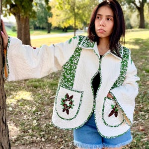 May include: A white quilted jacket with green leaf pattern trim and pockets. The jacket features embroidered pinecone and berry designs on the pockets. The sleeves are flared, and the jacket is open in the front.