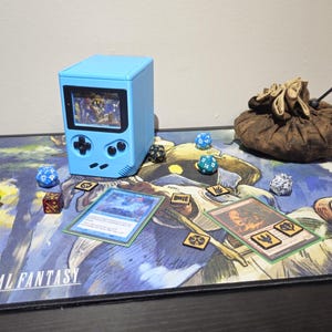 May include: A blue gaming console-shaped box with a screen, dice, cards, and a brown drawstring bag are on a Magic: The Gathering playmat. The playmat features a Final Fantasy design. Various dice are scattered around the cards.