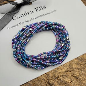 May include: A stack of beaded bracelets in shades of blue, purple, and silver. The bracelets are made of small, round beads and feature geometric silver accents. The bracelets are on a white surface with the text "Candra Ella Custom Handmade Beaded Bracelets".