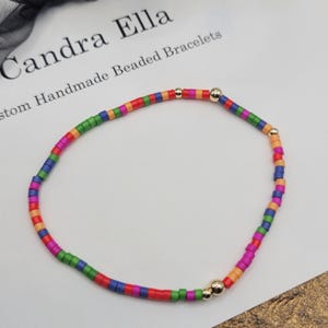 May include: A colorful beaded bracelet with small, rectangular beads in a rainbow of colors. The bracelet includes gold-colored accent beads and is displayed on a white surface. The text "Candra Ella Custom Handmade Beaded Bracelets" is visible in the background.