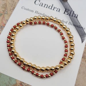 May include: Two beaded bracelets, one gold and one red, are stacked on top of each other. The bracelets are made of small, round beads.