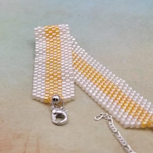 May include: A white and gold beaded bracelet with a silver clasp. The bracelet is made with small, square beads and has a simple, elegant design.