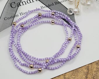 May include: Four beaded bracelets in shades of lavender and white, with gold-colored beads interspersed throughout. The bracelets are arranged in a circular pattern. The text "Candra Ella" and "Custom Handmade Beaded Bracelet" is visible.