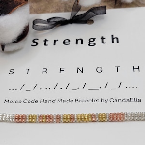 May include: A silver, gold, and rose gold bracelet with a woven design. The bracelet is labeled "Strength" in Morse code.